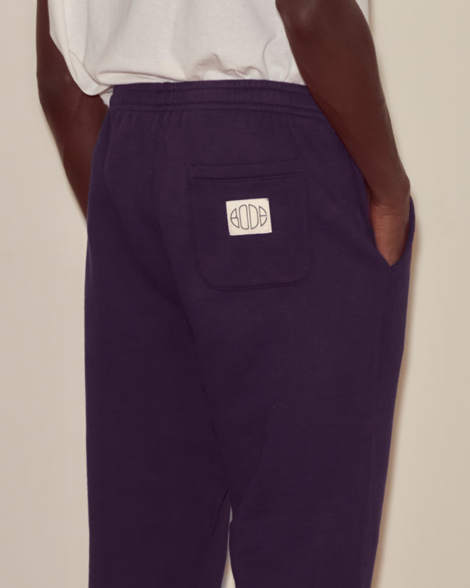 Person wearing Joggers - Dark Purple with a white patch on the back pocket, Bode woven label detail, hand in side pocket, and a white shirt showing.