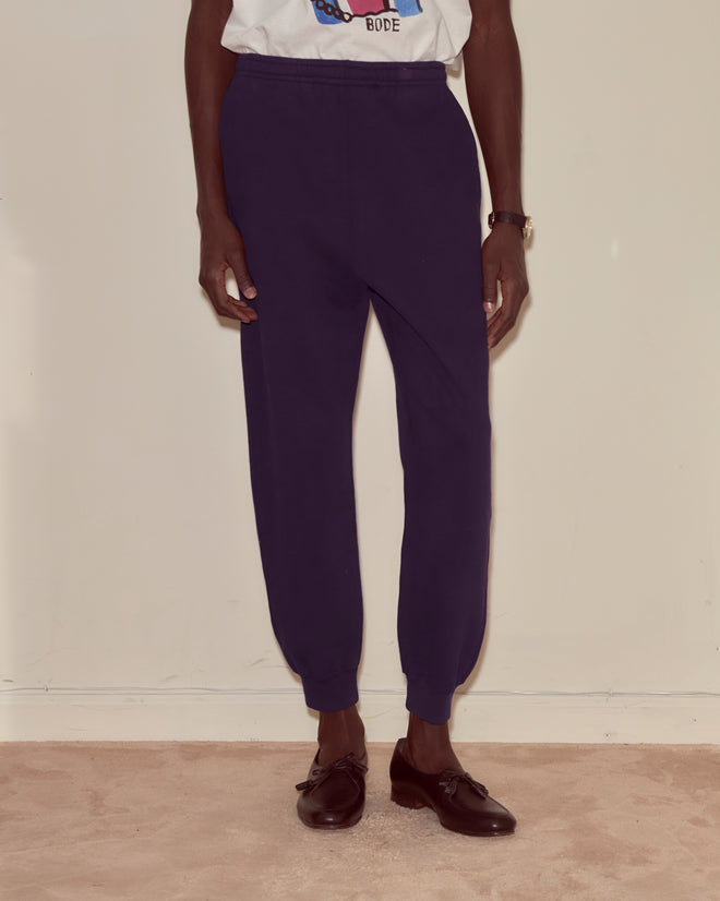 Person wearing dark purple Joggers with a Bode woven label, a white T-shirt, and black dress shoes stands on a beige carpet.