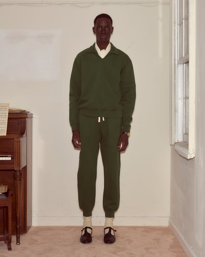 A person wearing the Crew Polo - Dark Green stands indoors by a piano and window, facing the camera.