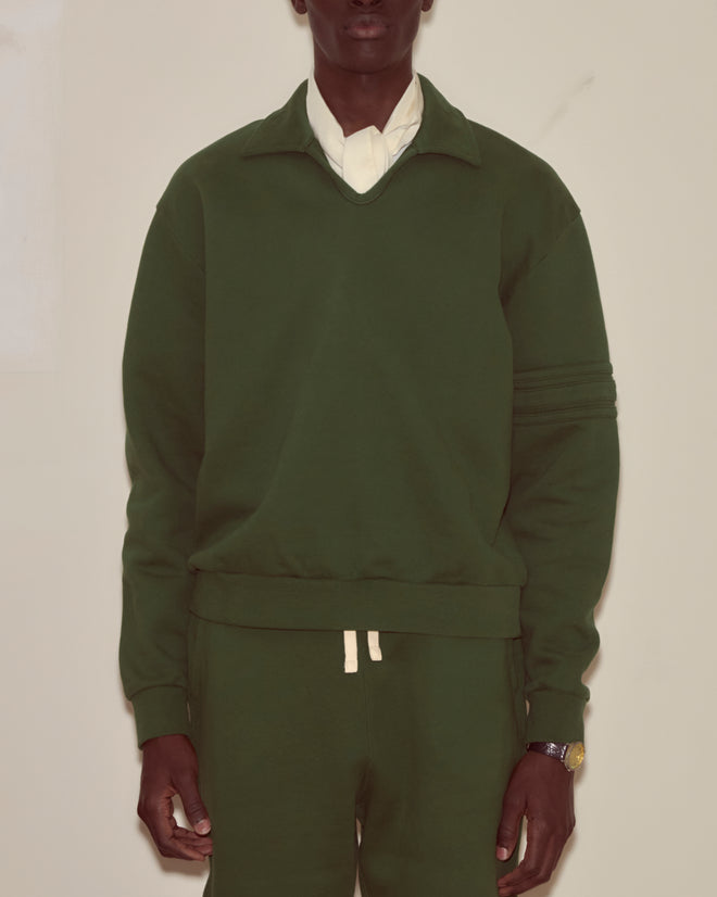 Wearing the Crew Polo - Dark Green, made from midweight cotton fleece and layered over a white shirt, a person stands against a light background.