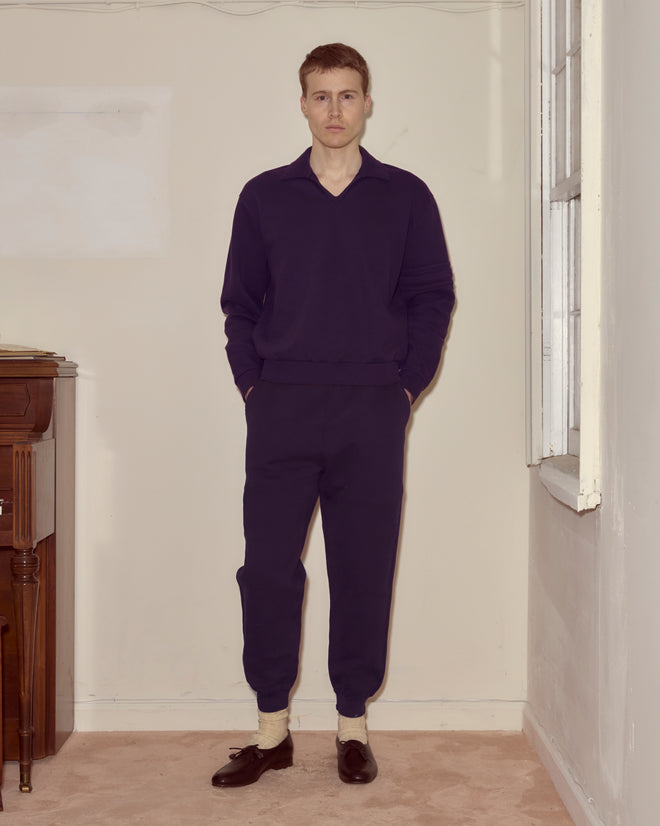 A man stands indoors wearing the Crew Polo in dark purple with his hands in his pockets.