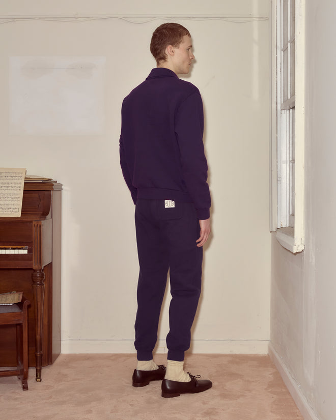 A man in a dark purple Crew Polo stands facing a window in a beige room, with a piano on the left.