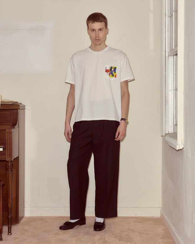 A man stands indoors wearing the Portrait Pocket Tee in white, paired with black pants and shoes with white socks—his clean-lined, understated look evokes modernist art.