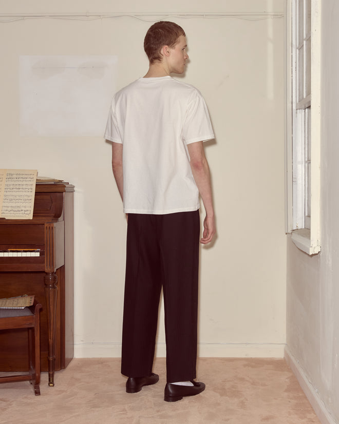 Wearing the Portrait Pocket Tee and black pants, a person stands facing a window in a minimalist room with a piano, reflecting Modernist art’s clean lines.
