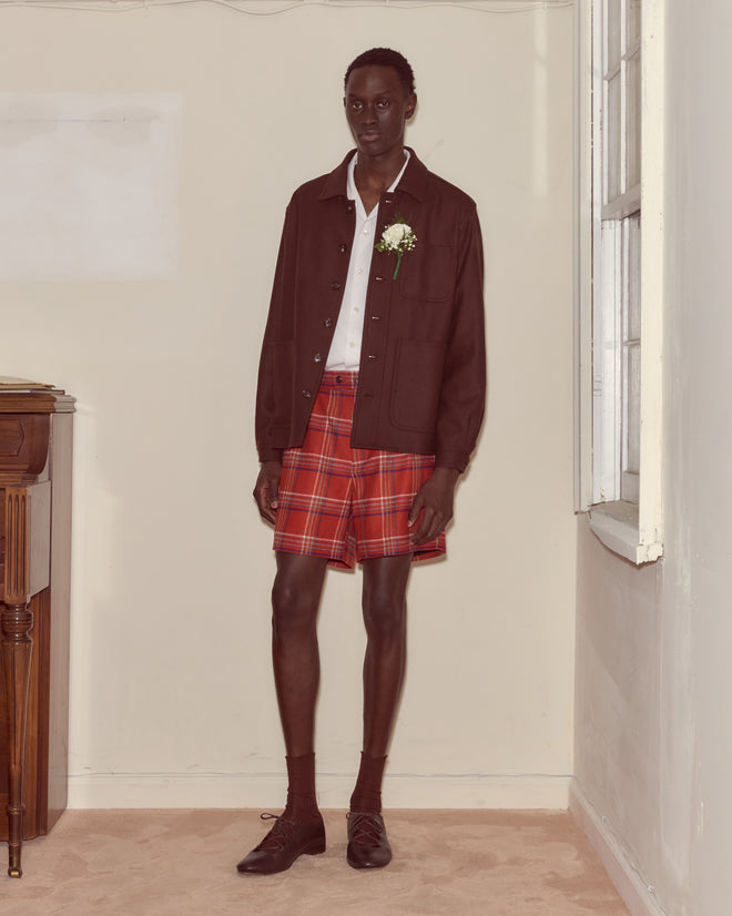Wearing the Bramble Franck Jacket with a white shirt, red plaid shorts, and dress shoes, a person stands indoors by a window, blending classic suiting with a touch of 1960s workwear style.