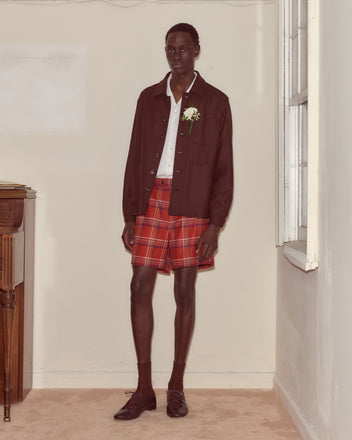 A person stands indoors wearing the Bramble Franck Jacket styled with a white shirt, red plaid shorts, socks, and dress shoes.