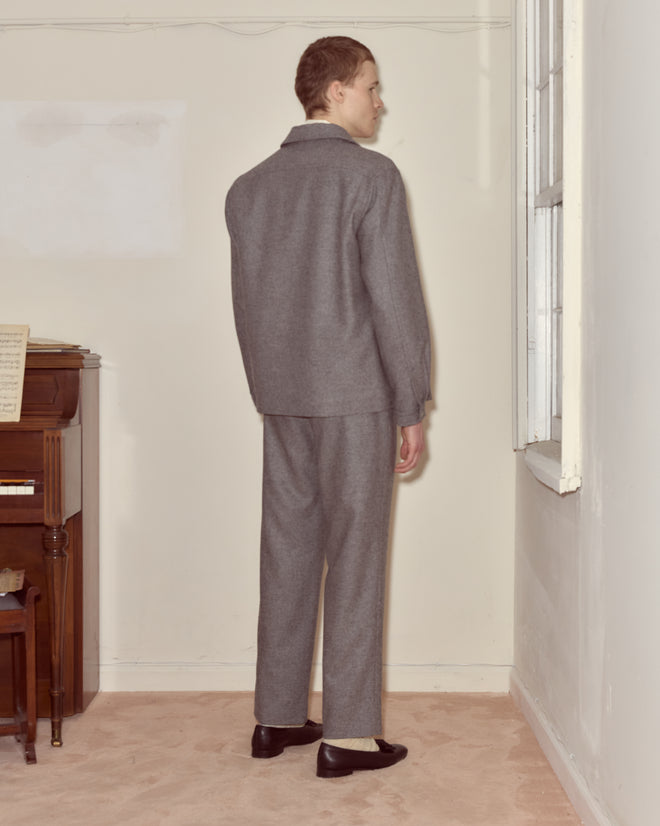 A man wearing the Dovecote Franck Jacket in heathered grey stands facing a window in a sparsely furnished room with beige carpet.