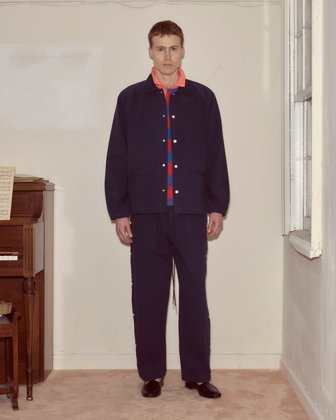 A man stands indoors by a piano and window, wearing the Twill Track Jacket - Navy over a coral shirt with matching pants.