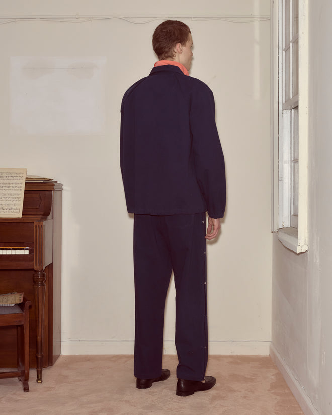 A man in a navy Twill Track Jacket stands facing the corner of a sparsely furnished room with a piano on the left.