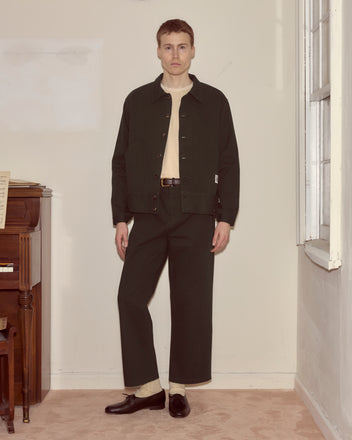A man stands indoors by a piano and window, wearing dark pants and a Twill Knolly Brook Side Tab Jacket in Dark Green over a light shirt.