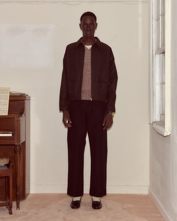 Indoors by a piano, a person wears dark pants, light shoes, and the Waxed Coach's Jacket in Brown.