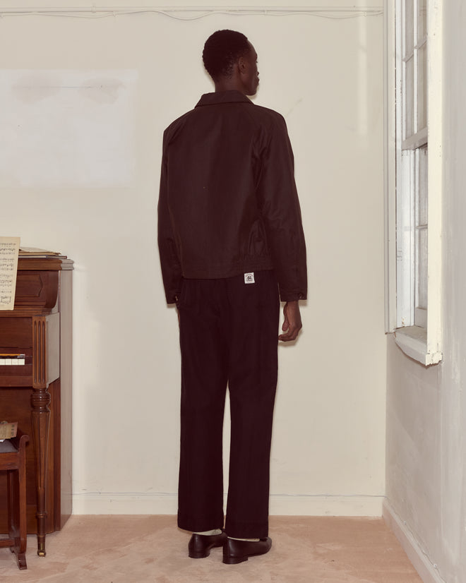 A person wearing the Waxed Coach's Jacket in brown stands facing a corner in a room with a piano and sheet music to the left.