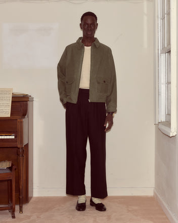 A person in a Waxed Coach's Jacket - Light Green, cream shirt, black pants, and loafers stands indoors near a piano and window.