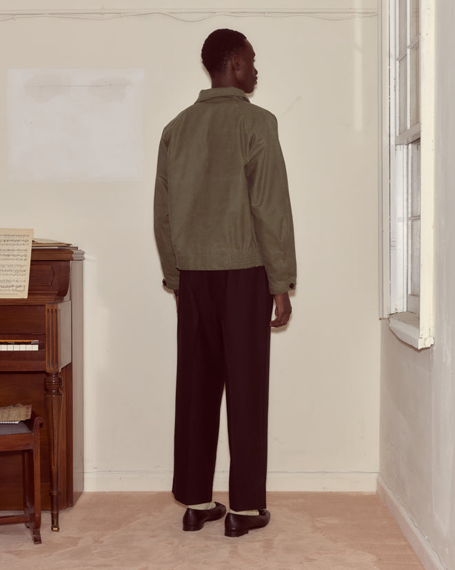 A person in a Waxed Coach's Jacket - Light Green and black pants stands facing a cream wall near a piano and window.
