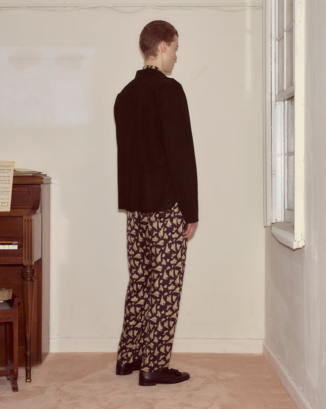 A person stands indoors facing a corner, styled in patterned pants and the Suede Overshirt - Black, next to a piano.
