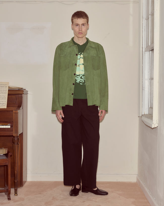 A person stands indoors beside a piano, wearing the Suede Overshirt - Green with black pants and shoes.