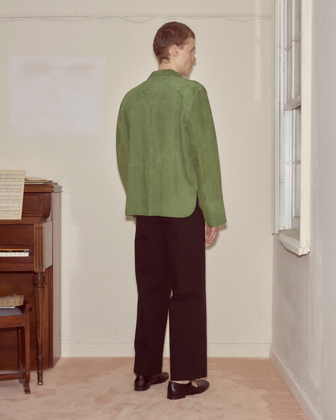 Person standing in a room facing away, wearing the Suede Overshirt - Green and black pants, near a piano and a window.