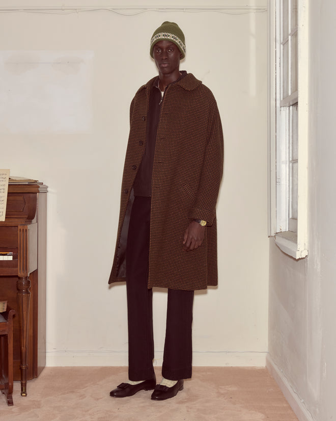 Wearing a green Moritz Hat, a person in a brown checked coat and black pants stands indoors near a piano and window, showcasing luxury accessories.