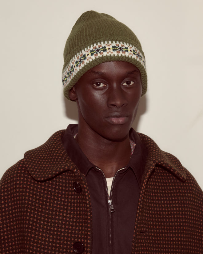 Wearing the green Moritz Hat, crafted from soft merino wool, and a brown patterned coat, the person gazes at the camera with a neutral expression, embodying understated luxury accessories style.