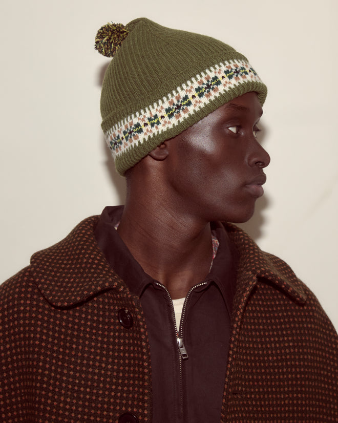 A man in a green Moritz Hat and a brown patterned coat looks to the right against a plain background.