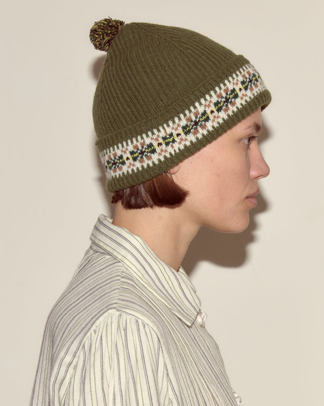 A person in profile wears the Moritz Hat, a green Fair Isle merino wool accessory with a patterned band, paired with a striped button-up shirt for a cozy, luxurious look.