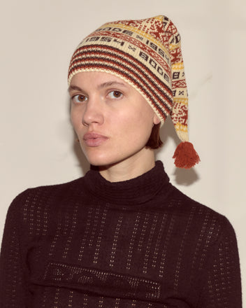 A person wearing the Marzipan Hat, featuring a Norwegian pattern, 100% wool construction, and a tassel, paired with a dark textured sweater, looks at the camera.