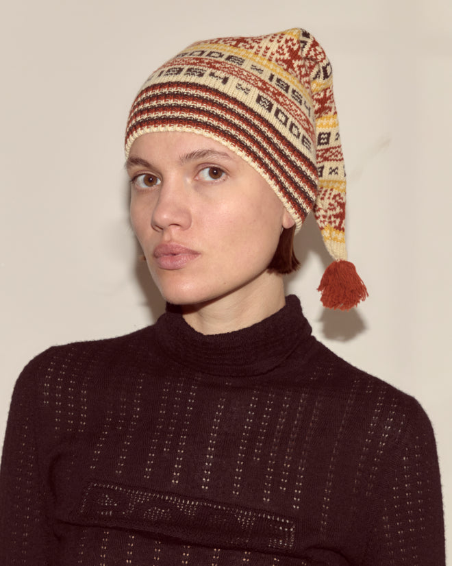 A person wearing the Marzipan Hat, featuring a Norwegian pattern, 100% wool construction, and a tassel, paired with a dark textured sweater, looks at the camera.