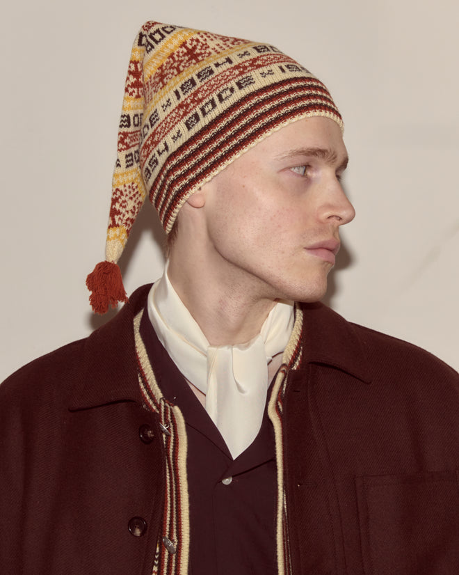 Person in a Marzipan Hat, brown Bode coat, and white scarf looks to the side against a plain background.