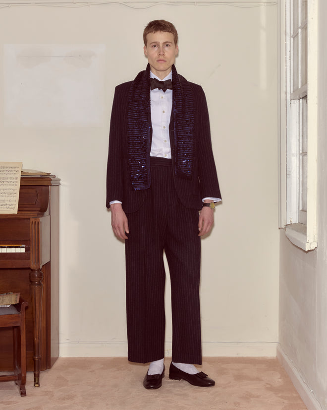 A person in a tuxedo pairs the Sequin Spotlight Scarf - Black, standing indoors beside a piano and facing the camera.