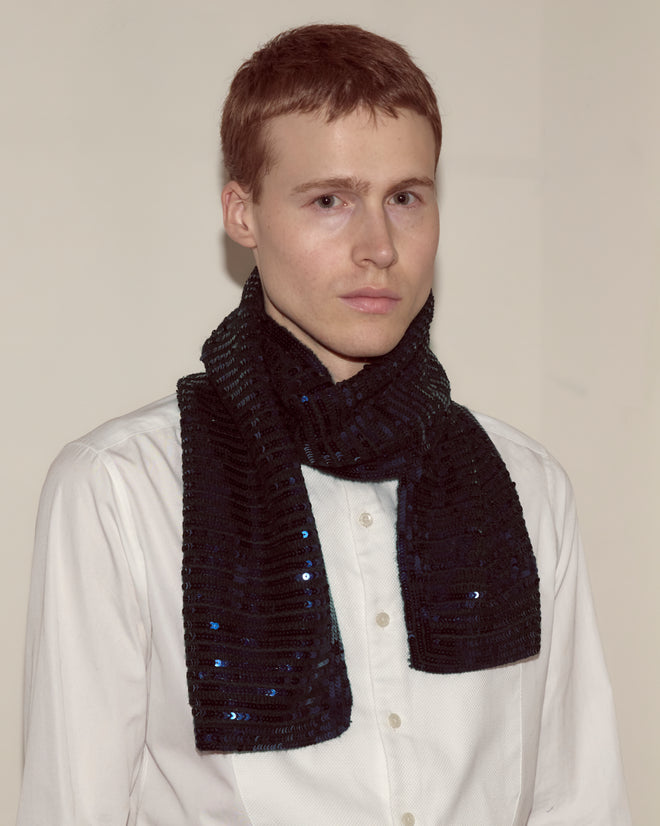 A person in a white shirt wears the Sequin Spotlight Scarf - Black, one of the season's standout knit accessories, and looks at the camera with a neutral expression.