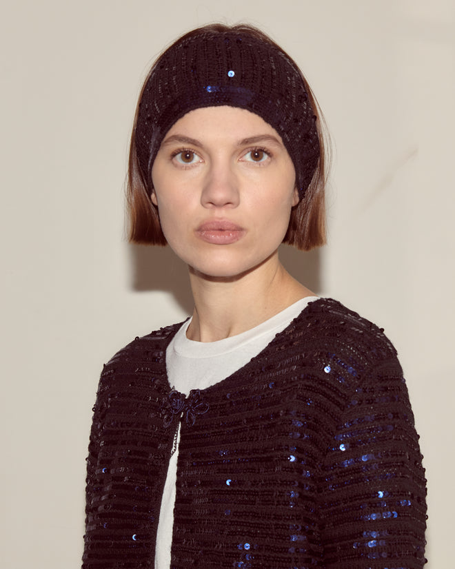 A woman wears the Sequin Spotlight Headband—this season's must-have knit accessory—with a matching jacket and white top, looking at the camera.