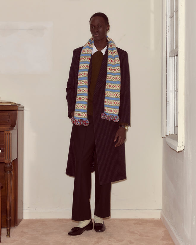 A person in a long dark coat and a Wedgewood Scarf stands in a corner by a window and a wooden piano.