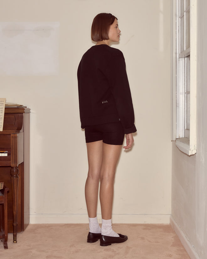 Woman in a Double-Breasted Cardigan - Black stands sideways near a window, facing away from the camera.