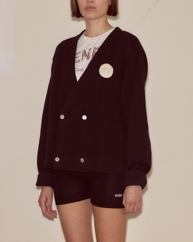 Woman in a Double-Breasted Cardigan - Black, paired with black shorts and standing against a plain wall.