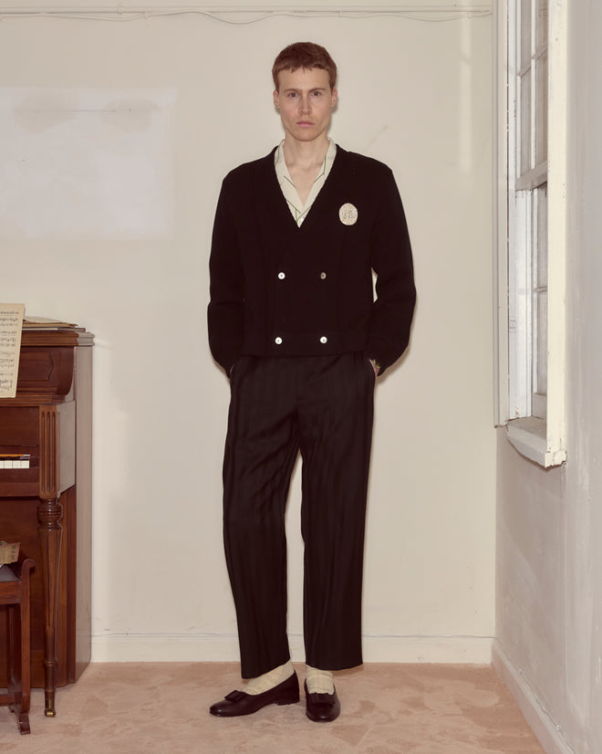 A man in a Double-Breasted Cardigan - Black, white shirt, and black pants stands in a room by a window and a piano.