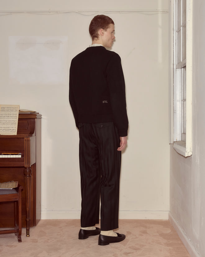 A person wearing the Double-Breasted Cardigan - Black stands facing a wall in a room with a piano and sheet music.