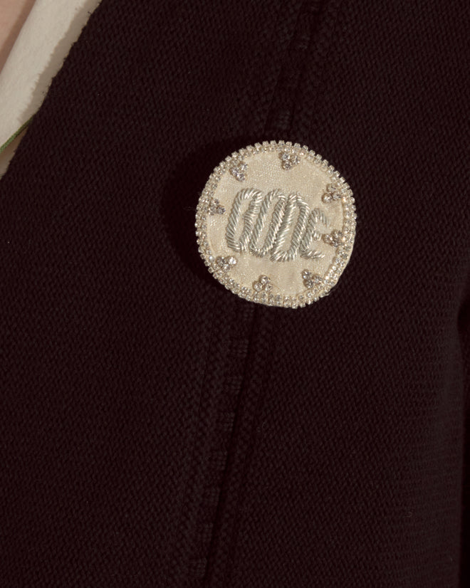 A round white embroidered patch with silver accents elevates the Double-Breasted Cardigan - Black, adding a refined touch to its textured knitwear design.