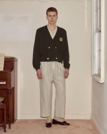 A man in striped pajamas, a dark green double-breasted cardigan, and loafers stands near a piano in a softly lit room.