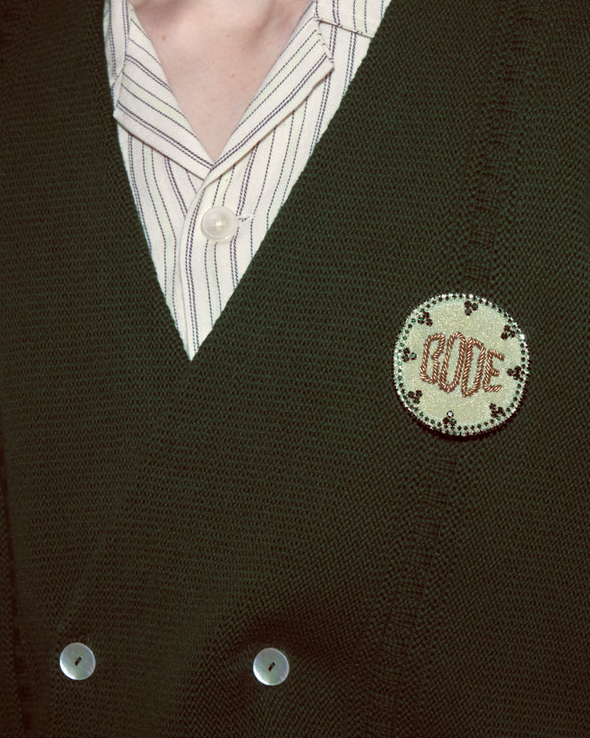 A person wearing a Double-Breasted Cardigan - Dark Green with a round "GOOD" pin and a striped shirt underneath.