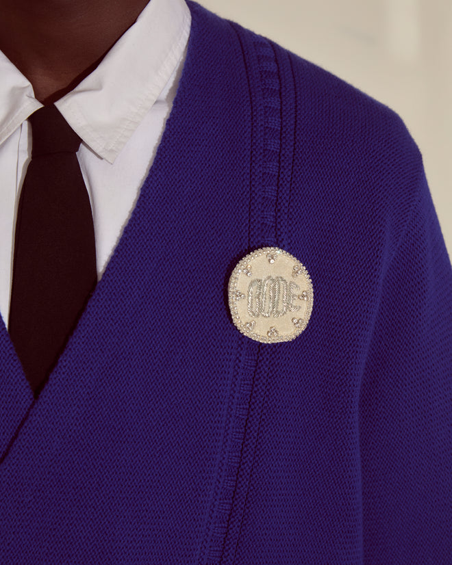 Person wearing a Double-Breasted Cardigan - Ultramarine by Bode, white shirt, black tie, and a round jeweled brooch with 