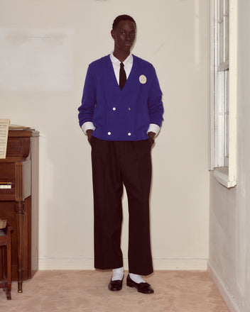 Wearing the Double-Breasted Cardigan - Ultramarine with black pants and a tie, a person stands indoors by a piano and window, hands in pockets, capturing the timeless 1960s golf cardigan look.