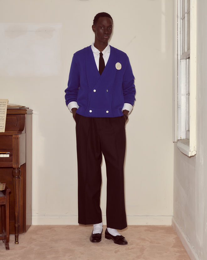 Wearing the Double-Breasted Cardigan - Ultramarine with black pants and a tie, a person stands indoors by a piano and window, hands in pockets, capturing the timeless 1960s golf cardigan look.
