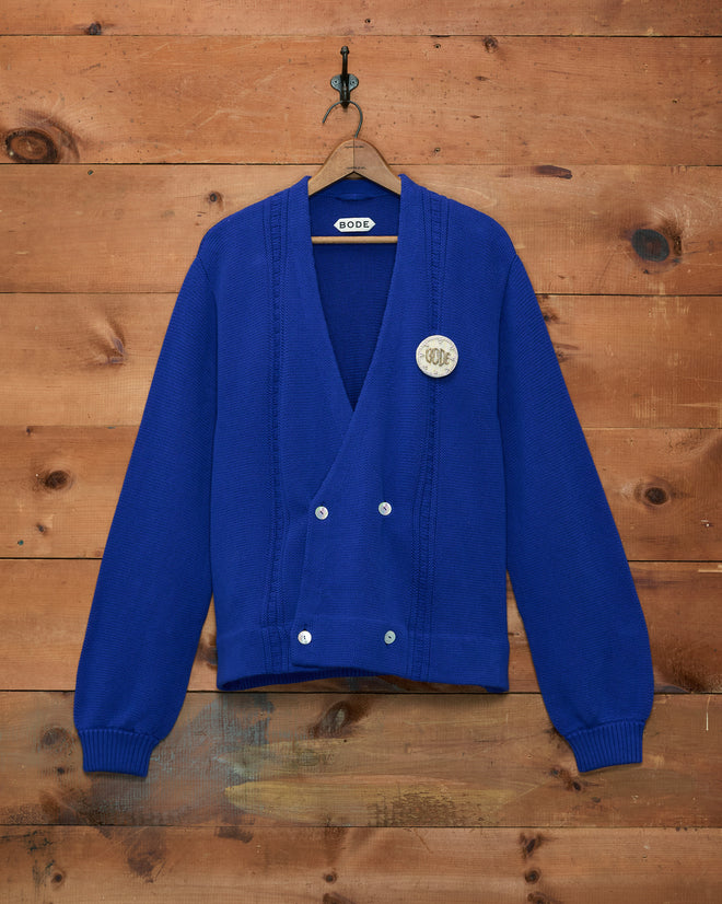 The Double-Breasted Cardigan - Ultramarine, featuring silver buttons and a round white hand-embroidered patch for a unique touch, hangs on a wooden wall.