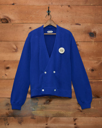 The Double-Breasted Cardigan - Ultramarine, featuring silver buttons and a round white hand-embroidered patch for a unique touch, hangs on a wooden wall.