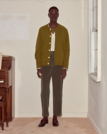 A person stands indoors wearing the Orchard Cardigan in green, paired with a white shirt, green pants, and brown shoes.