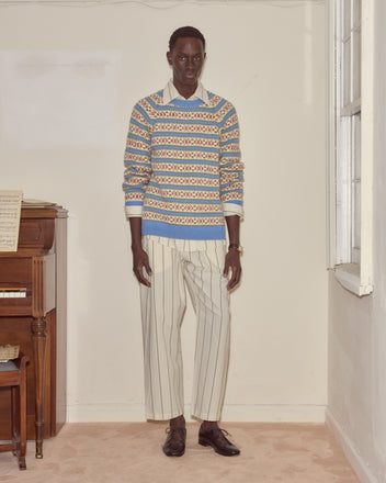 Wearing the Wedgewood Sweater and striped pants, a person stands indoors by a piano and window.