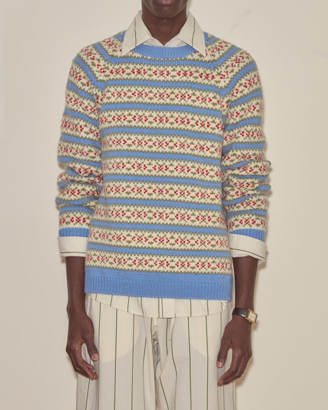 A person wears the Wedgewood Sweater, a vintage knitwear piece with pastel patterns, over a collared striped shirt and matching striped pants.