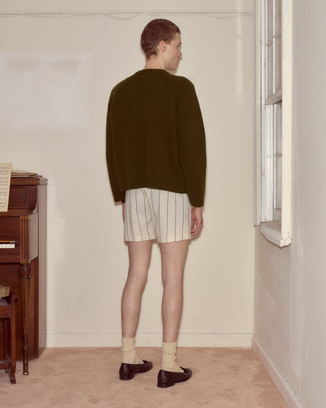 Wearing a boxy silhouette Cashmere Crewneck - Hemlock sweater, striped shorts, socks, and black shoes, a person stands facing a wall in a sparse room.