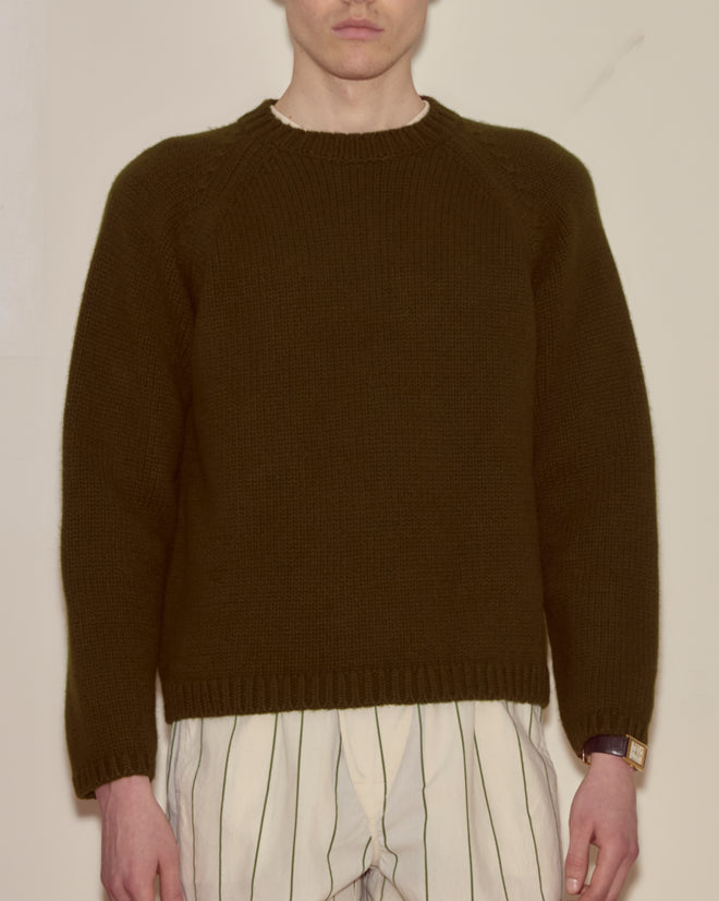 A person wearing the Cashmere Crewneck - Hemlock, a dark green sweater, paired with white pants featuring black vertical stripes, stands against a plain wall.