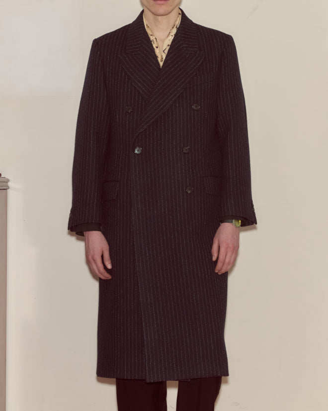 A person wears the Waldorf Pinstripe Coat—a long, navy wool coat—over a collared shirt, standing against a plain background.